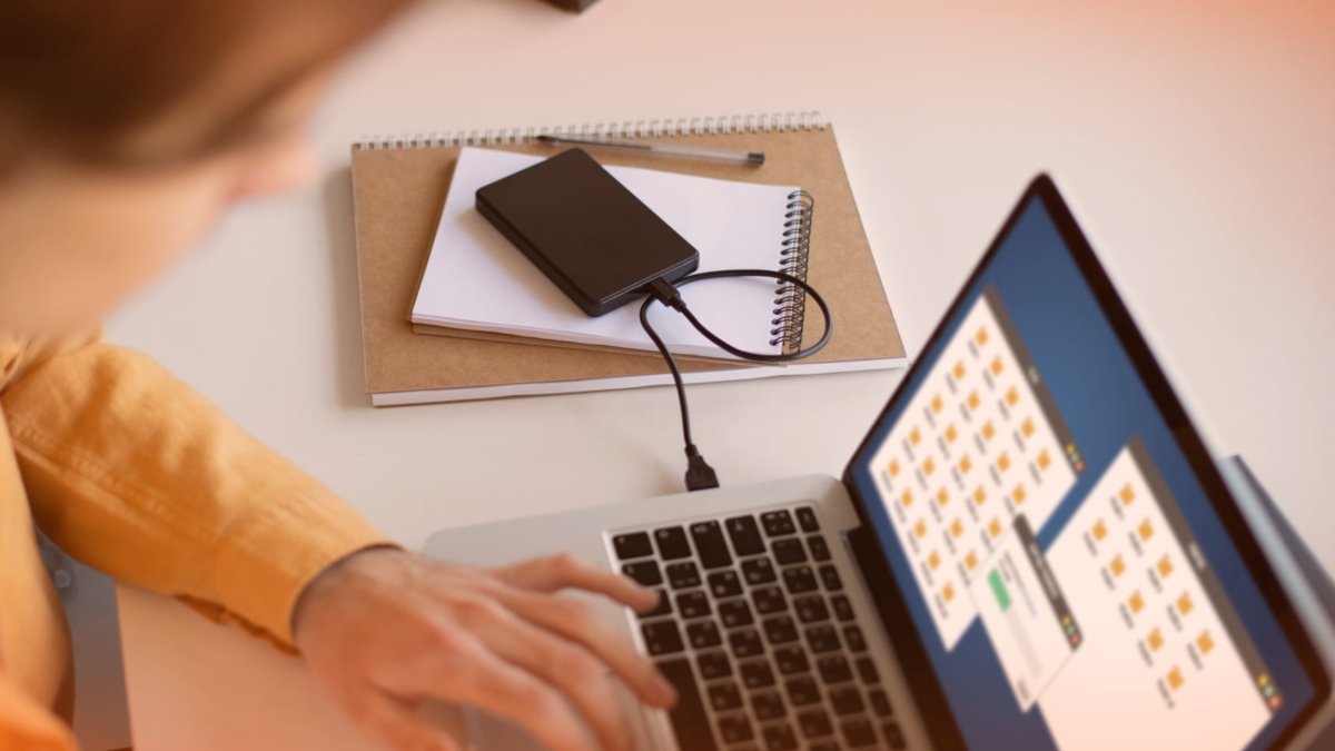 Person using a laptop with external hard drive and notebooks, illustrating IT support and data management in a professional setting.